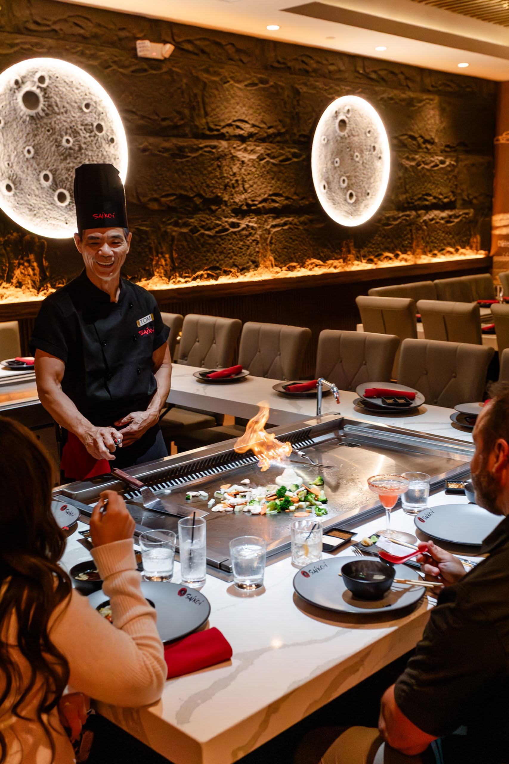 A hibachi chef cooking vegetables on a large flat-top grill with a small flame for seated guests at Saiko-i.