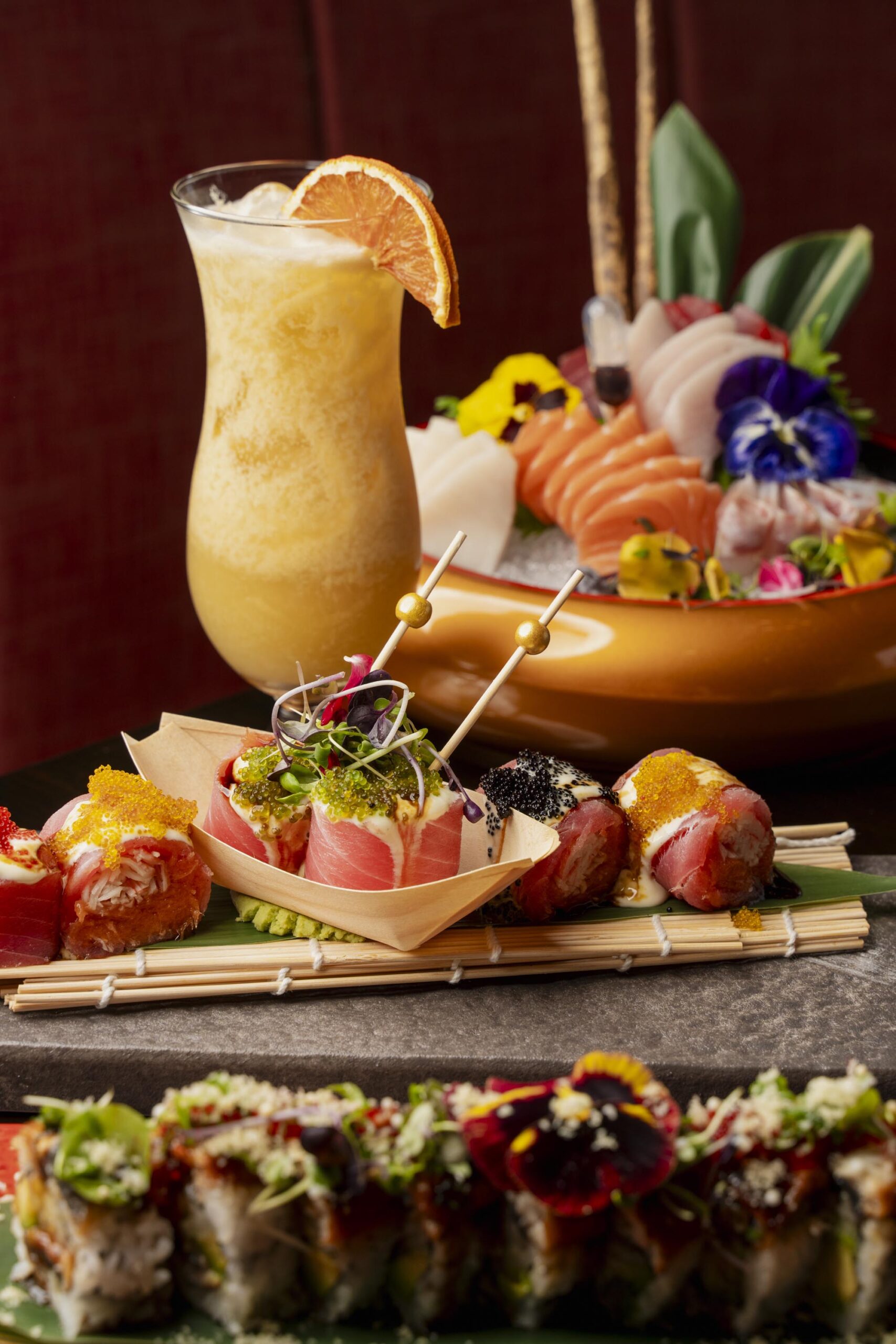 Variety of specialty sushi rolls garnished with fish roe and edible flowers, served with a frozen tropical cocktail.